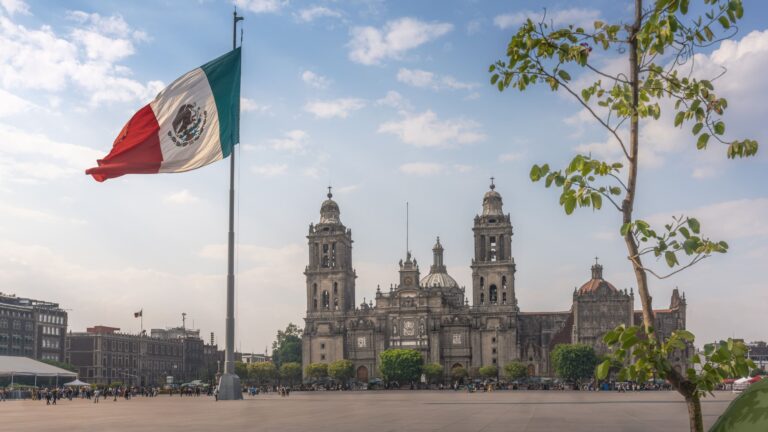Zócalo Square in Mexico City, with the Mexican national flag flying in the foreground and the Metropolitan Cathedral rising behind it under an open sky.