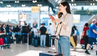 A young Asian woman standing in an international airport departure terminal, looking at her smartphone while carrying travel luggage.