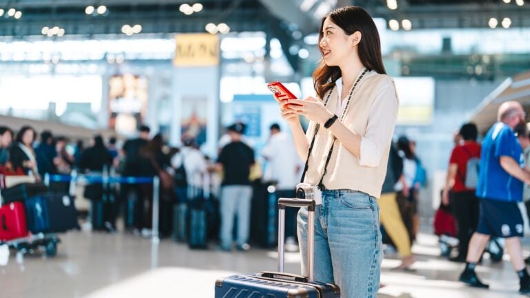 A young Asian woman standing in an international airport departure terminal, looking at her smartphone while carrying travel luggage.