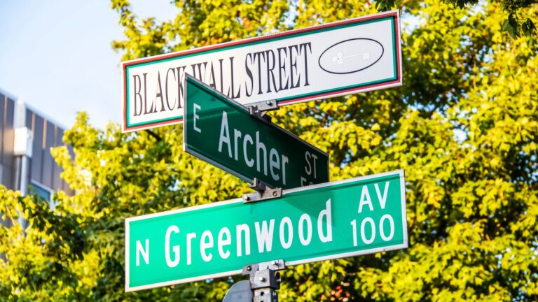 Close-up of street signs for Black Wall Street and N. Greenwood Avenue at the Archer Street intersection in Tulsa, Oklahoma, with a blurred bokeh background.