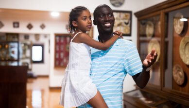 A smiling Black father and his young daughter viewing an exhibition together inside a museum.