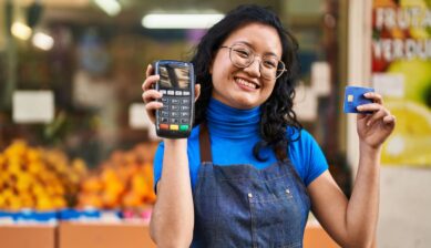 A young Chinese woman working at a fruit store holds a credit card reader and a credit card, ready to process a mobile payment transaction.