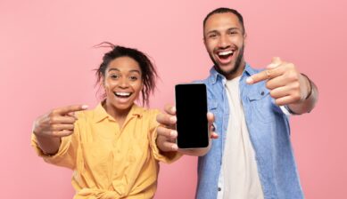 A smiling Black couple outdoors, both pointing enthusiastically at a smartphone screen displaying a blank screen, suggesting a mobile app or website recommendation.