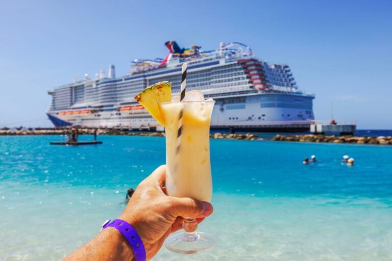 Close-up view of hand holding glass of cocktail on Caribbean beach with a cruise in background.