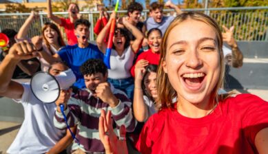A group of diverse, smiling fans taking a selfie together in sunny stadium stands, visibly celebrating during a football match.