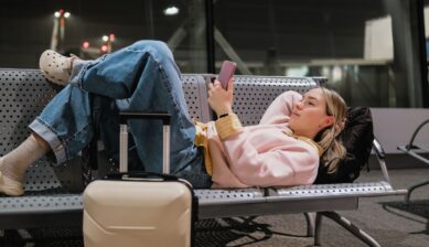A young woman sitting alone on an airport bench at night, luggage beside her and smartphone in hand, waiting for a delayed flight.