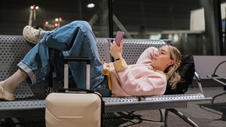 A young woman sitting alone on an airport bench at night, luggage beside her and smartphone in hand, waiting for a delayed flight.