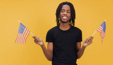 A smiling young man holding two small American flags against a yellow background, celebrating the Juneteenth national holiday.
