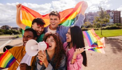 A diverse group of young activists smiling and cheering while marching in an LGBTQ+ Pride parade, holding rainbow flags and megaphones in an urban setting.
