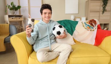 A teenage boy holding a Mexican flag and snacks watching a soccer match, expressing excitement as a fan at home during a major tournament.