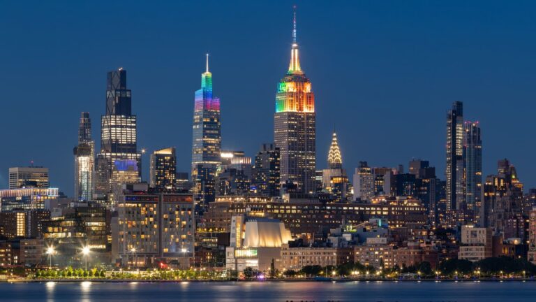 Midtown Manhattan skyscrapers illuminated with rainbow colors for Pride Week, viewed from across the Hudson River at dusk.