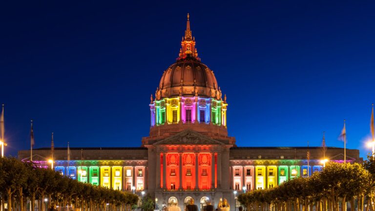 San Francisco City Hall lit up in rainbow colors at night during Pride Week, with the illuminated dome reflected against the dark sky.