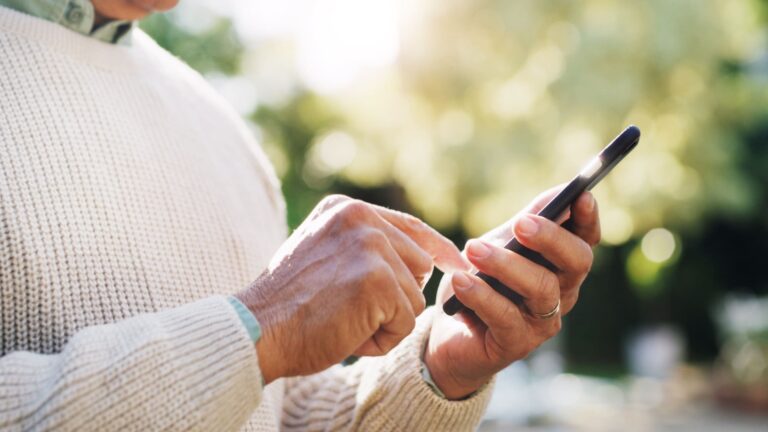 An elderly man sitting outdoors scrolling on a smartphone, appearing to browse or download a mobile application.