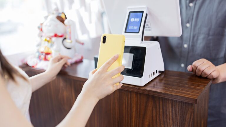 A cashier holds a smartphone displaying a QR code while a customer pays at a café counter, using either Alipay or WeChat Pay for a contactless mobile transaction.