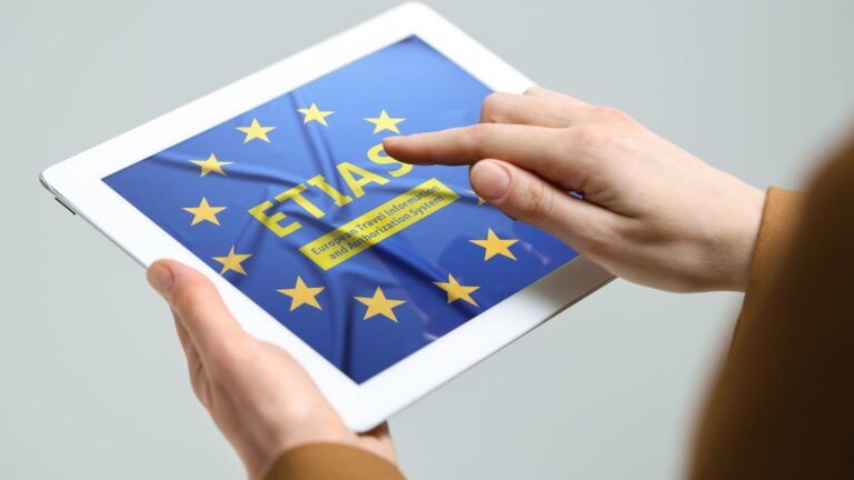 Woman holding a tablet displaying the ETIAS European Travel Information and Authorisation System webpage against a light grey background.