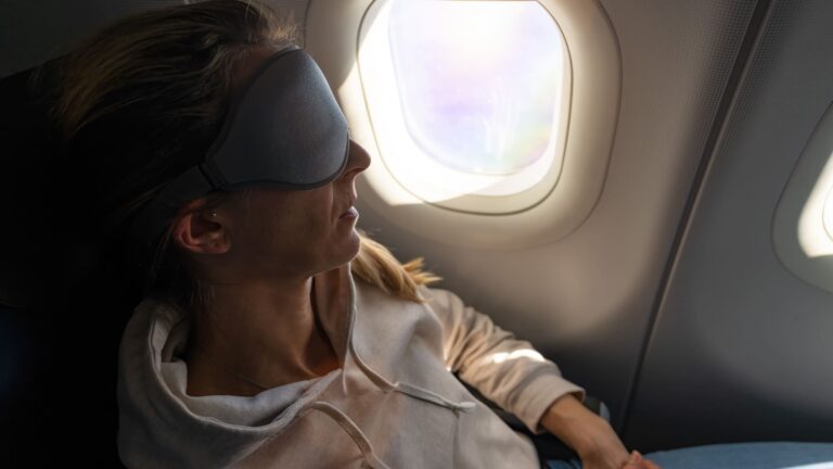 Woman wearing a sleep mask reclining in an airplane seat, with soft daylight coming through the cabin window beside her.