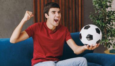 A young man in a red t-shirt holding a soccer ball, making a victory gesture while watching a live match on TV from his sofa at home.