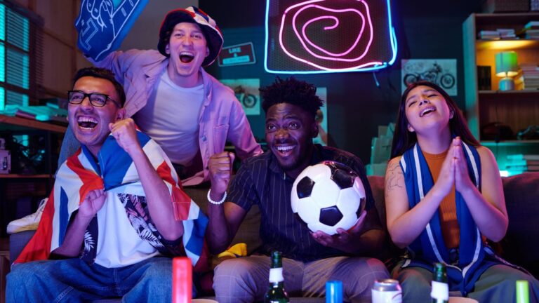A diverse group of young adults cheering and celebrating in front of a television, one man holding a soccer ball, neon lighting in the background.