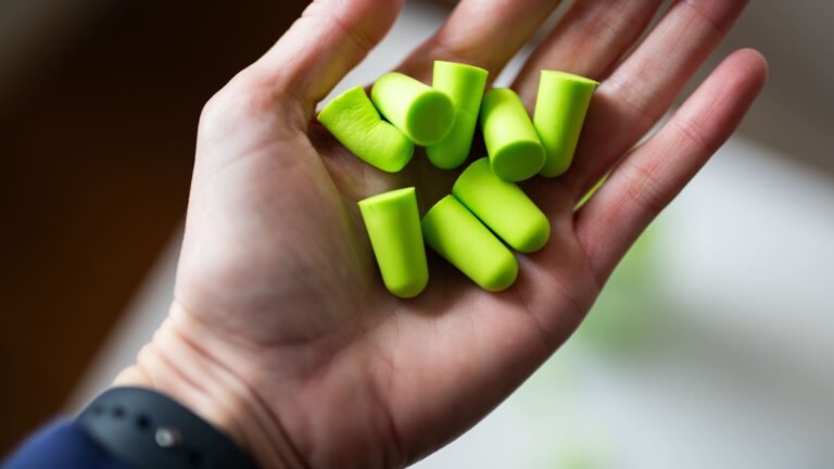 Hand holding green foam earplugs beside a jar against a white background