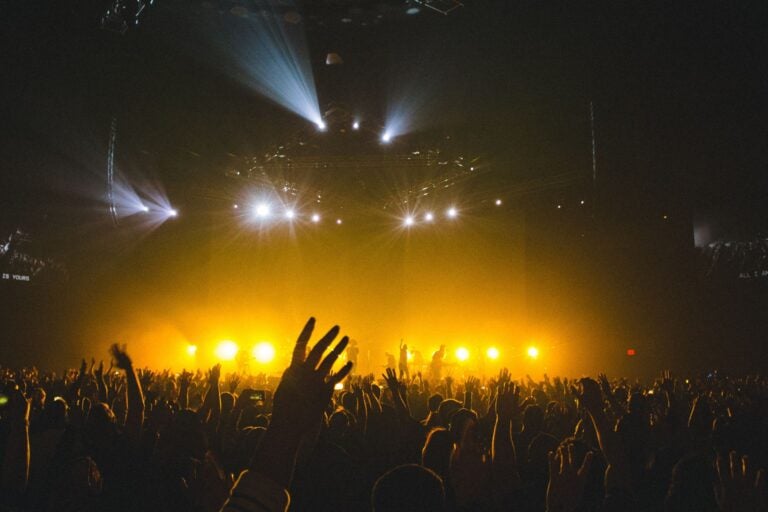 Silhouetted concert crowd with raised hands against warm stage lighting and spotlights.