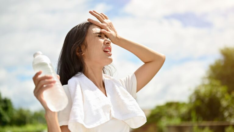 A young woman in lightweight sportswear pauses outdoors on a hot, sunny day, visibly overheated in the summer heat.