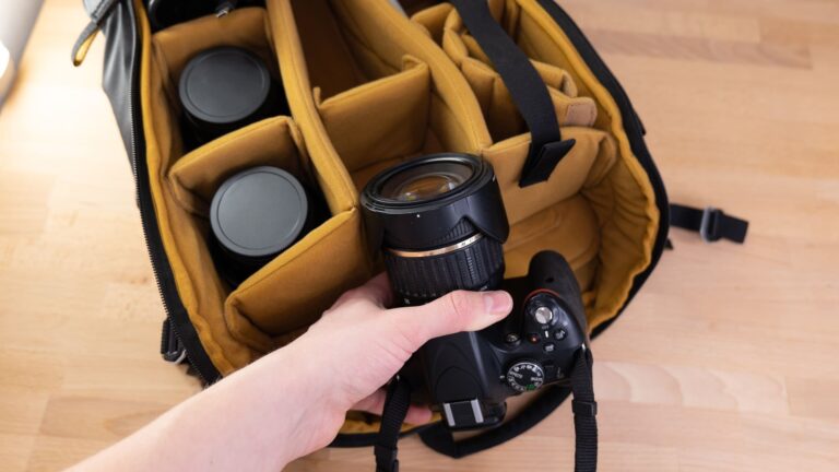 Person placing a camera lens into a backpack beside a camera on a wooden table