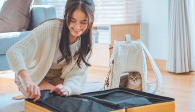 A young woman sits with her Scottish Fold cat while sorting clothes and packing them into an open suitcase before a trip.