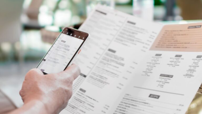A traveler seated at a restaurant table holds a menu in one hand and a phone running a translation app in the other.