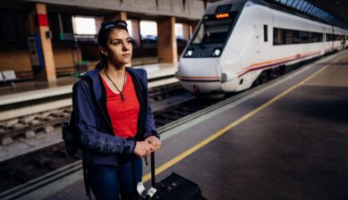 A tired traveler standing on a train station platform with a suitcase and bags, waiting for a train.