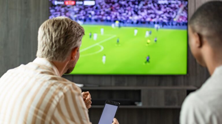 Two friends sitting at home in a living room, watching a football match while looking at a sports app on a smartphone.