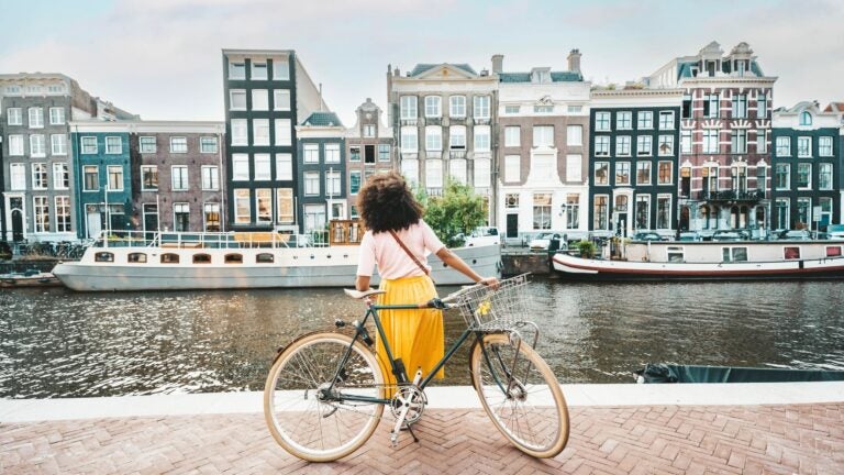 A smiling woman rides a bicycle along a city street in Amsterdam, with buildings and pedestrians in the background.