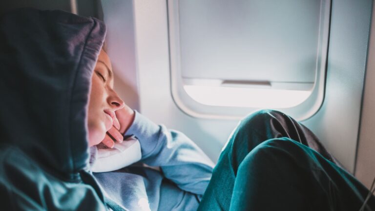 Blonde woman in a sporty hoodie asleep in an economy airplane seat, head tilted to one side during a flight.