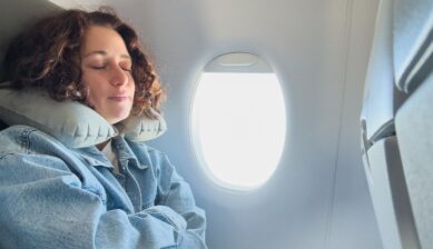 Young woman resting against an airplane window with a travel neck pillow, sleeping during a flight.