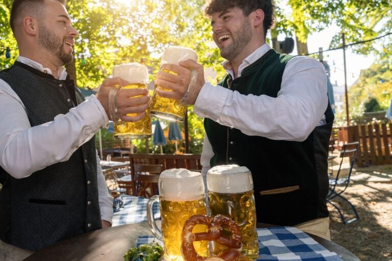 Two men in traditional Bavarian outfits toasting with large beer mugs at oktoberfest or beer garden.