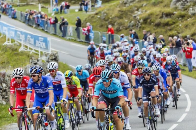 A packed peloton of road cyclists climbs a mountain road lined with spectators behind metal barriers.