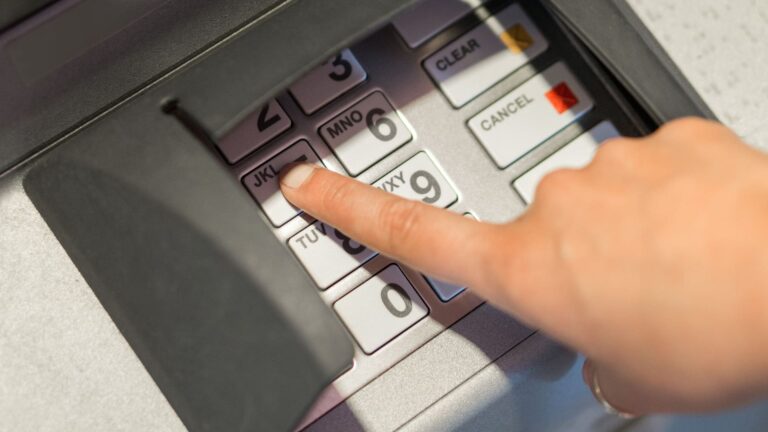 Close-up of a woman's hand entering a PIN on an ATM keypad while withdrawing cash from a machine.