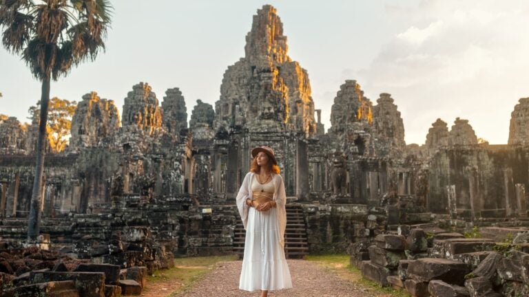 A woman in a white dress walks through the stone corridors of Bayon Temple at Angkor Wat, Cambodia, in golden-hour light.