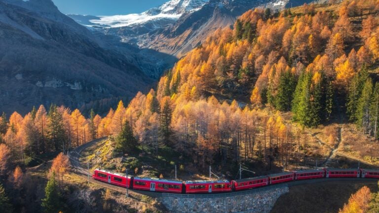 A red Bernina Express train winding along a curved mountain railway through an autumn forest in the Swiss Alps