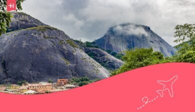 Scenic view of Olumo Rock in Abeokuta, Nigeria, with lush greenery and a cloudy sky.