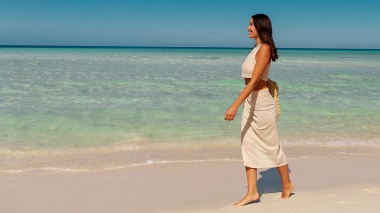 A woman carrying a bag walks along a sandy shoreline beside clear turquoise water, shown full length from the side.