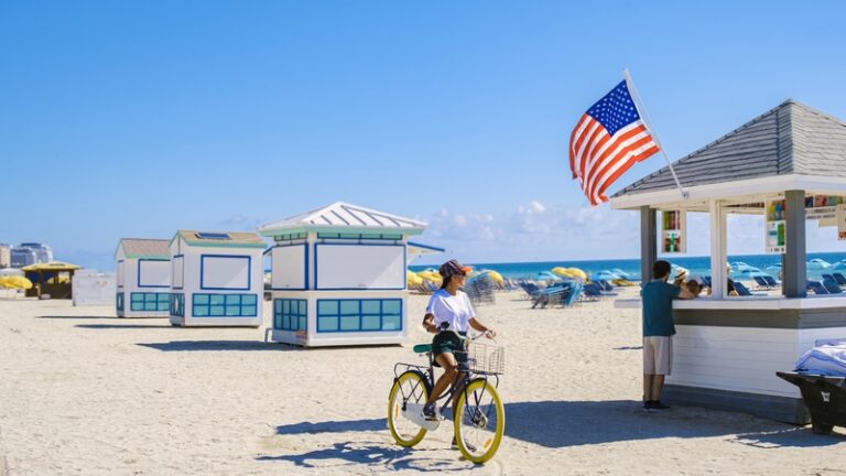A woman walking a bicycle past beach huts and a café on a sunny Florida beach.