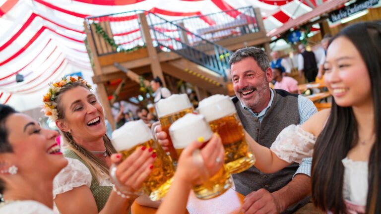 A group of friends laughing and clinking beer steins together at a long wooden table inside a red-and-white decorated Oktoberfest beer tent.