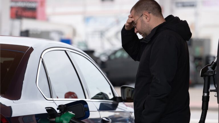 A frustrated man holding his head while standing at a gas station pump refueling his car.