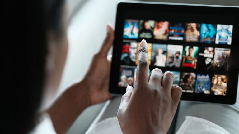 A woman lying on a sofa at home, browsing a streaming service app on her device to choose something to watch, shown from over her shoulder.
