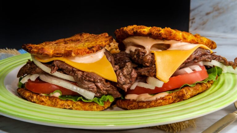 A jibarito sandwich using two flattened fried green plantains in place of bread, filled with steak, melted cheese, lettuce, and tomato, served with a side of rice and pigeon peas.