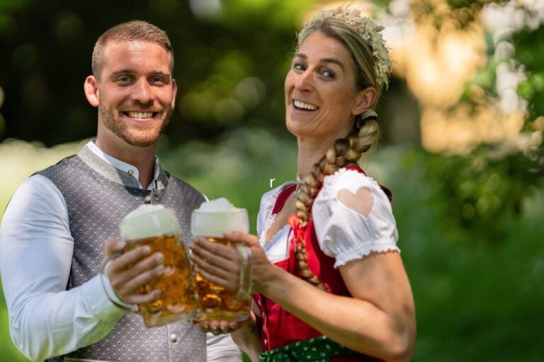 Happy Couple holding beer mugs and smiling, wearing a Dirndl and Lederhosen at Oktoberfest.