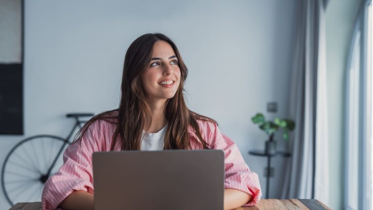 Young woman sitting at a desk with a laptop, pausing to look into the distance while planning