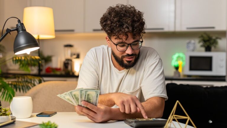 A man sits at a table using a calculator and taking notes while managing cash and monthly household expenses.