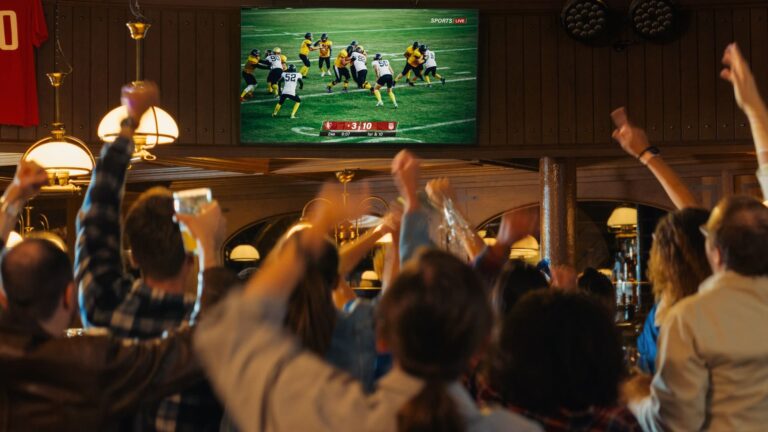A crowd of football fans in a sports bar cheering and celebrating as they watch a live match on a TV screen.