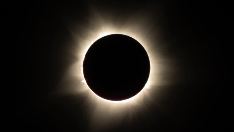 The sun's glowing white corona surrounds the completely darkened Moon against a black sky during the moment of totality